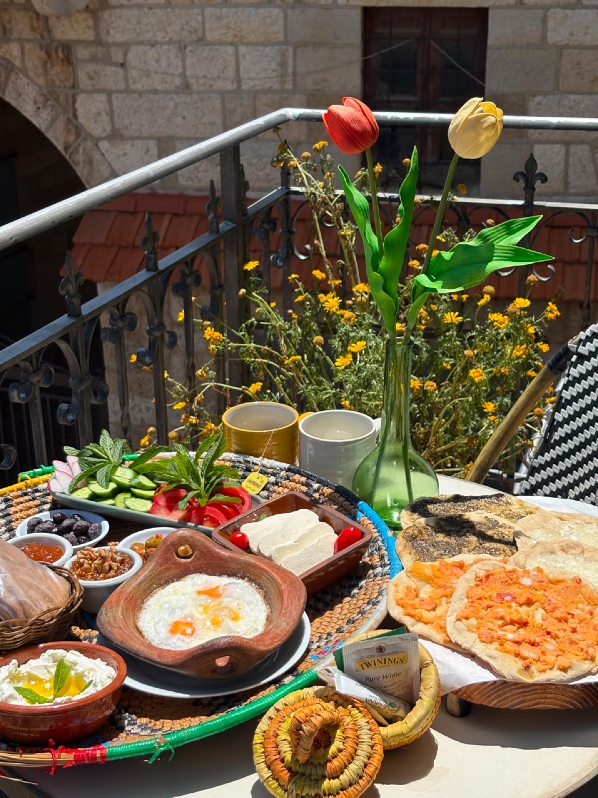 Lebanese village breakfast on the terrace — eggs in a clay pan, labneh, manousheh, fresh vegetables and tea, with the souk's stone walls behind