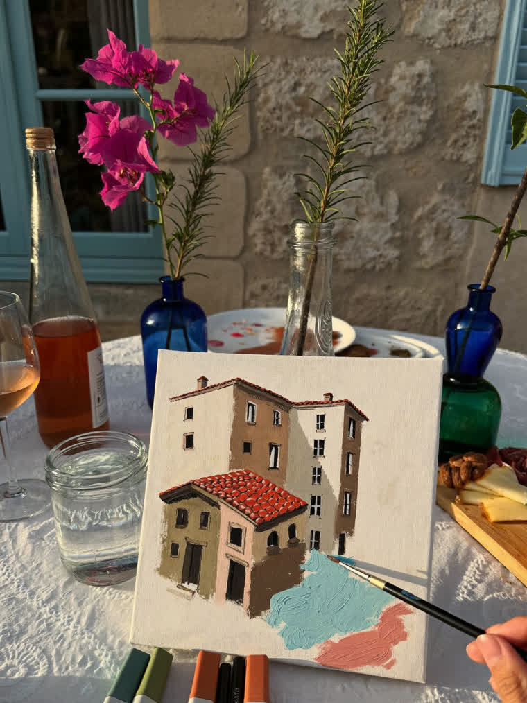 A small stretched canvas on the balcony table, half painted with a stone house and its red-tiled roof, beside rosé, rosemary in glass bottles and a cheese board