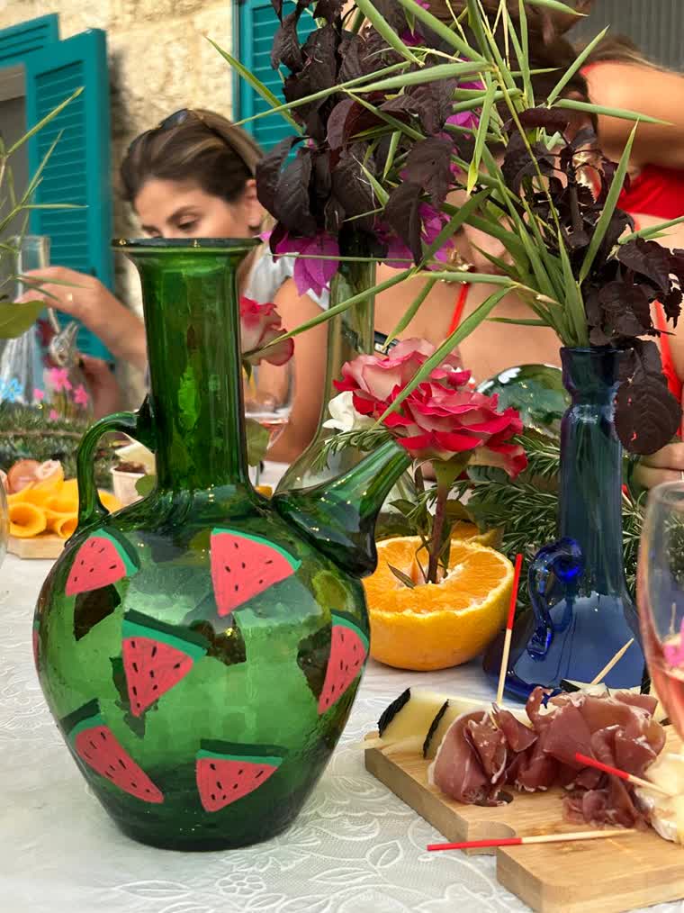 A green hand-blown glass ewer painted with pink watermelon slices, standing on the balcony table among flowers and a cheese board